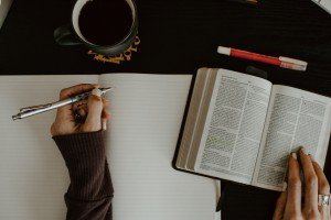 Hands writing in a notebook beside an open book and a cup of coffee