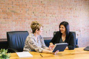 Two women talking across a meeting table, one holding a tablet