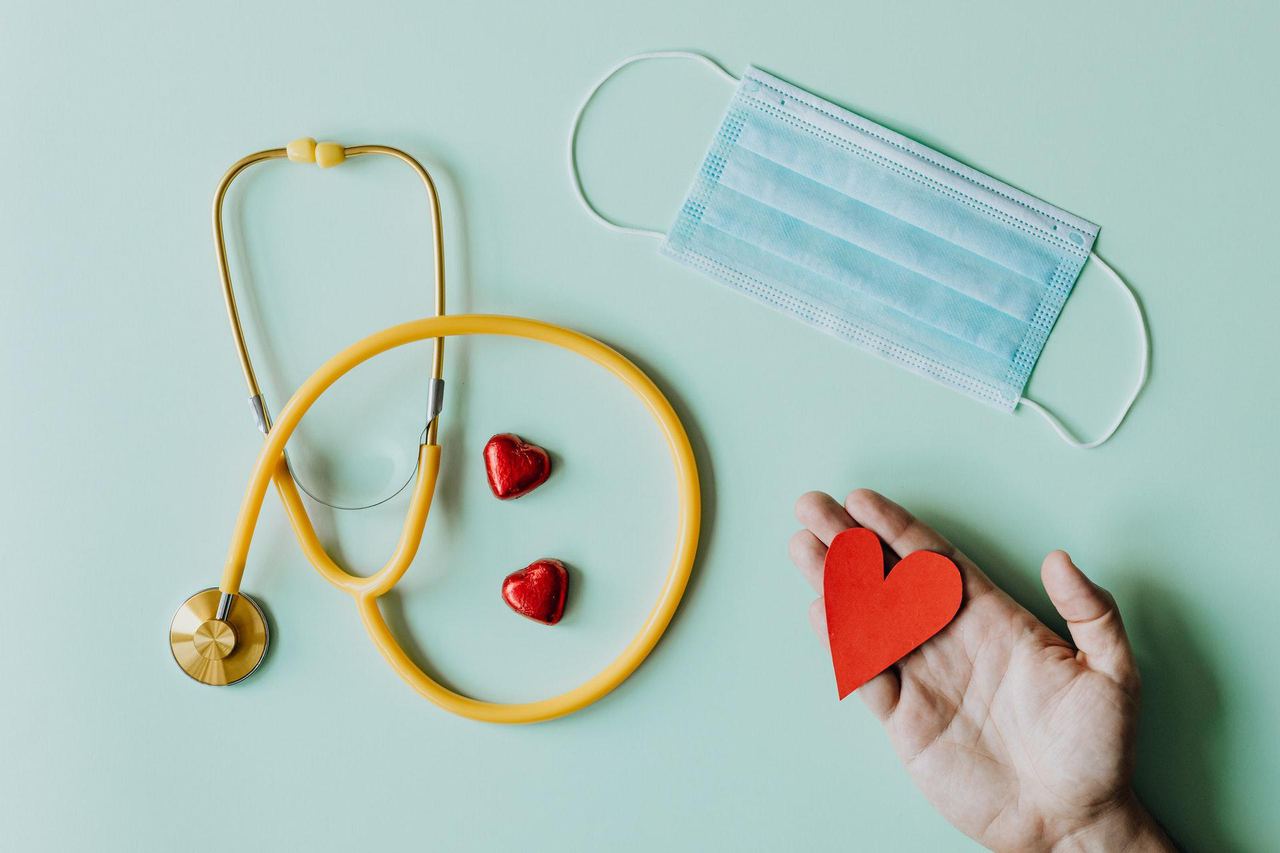 Stethoscope, face mask and red paper hearts on a pale green background